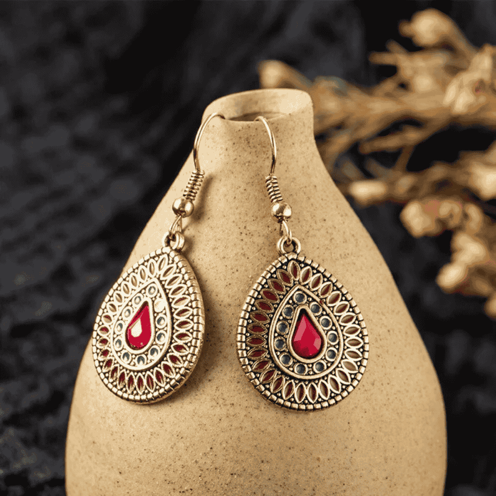 Close-up of a single antique gold teardrop drop earring with red and blue enamel on a light textured surface.