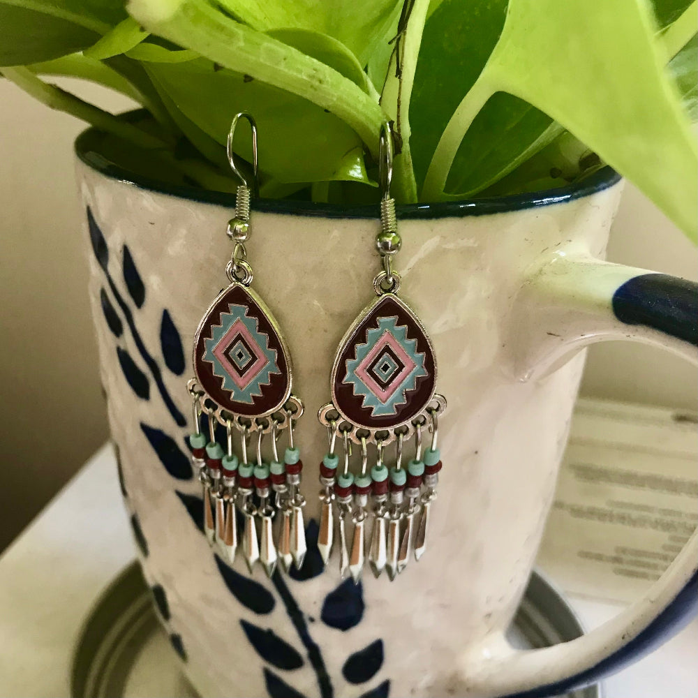 A pair of bohemian alloy teardrop dangle earrings with a geometric pattern in red, blue, and pink, hanging from a white and blue patterned mug with a green plant.