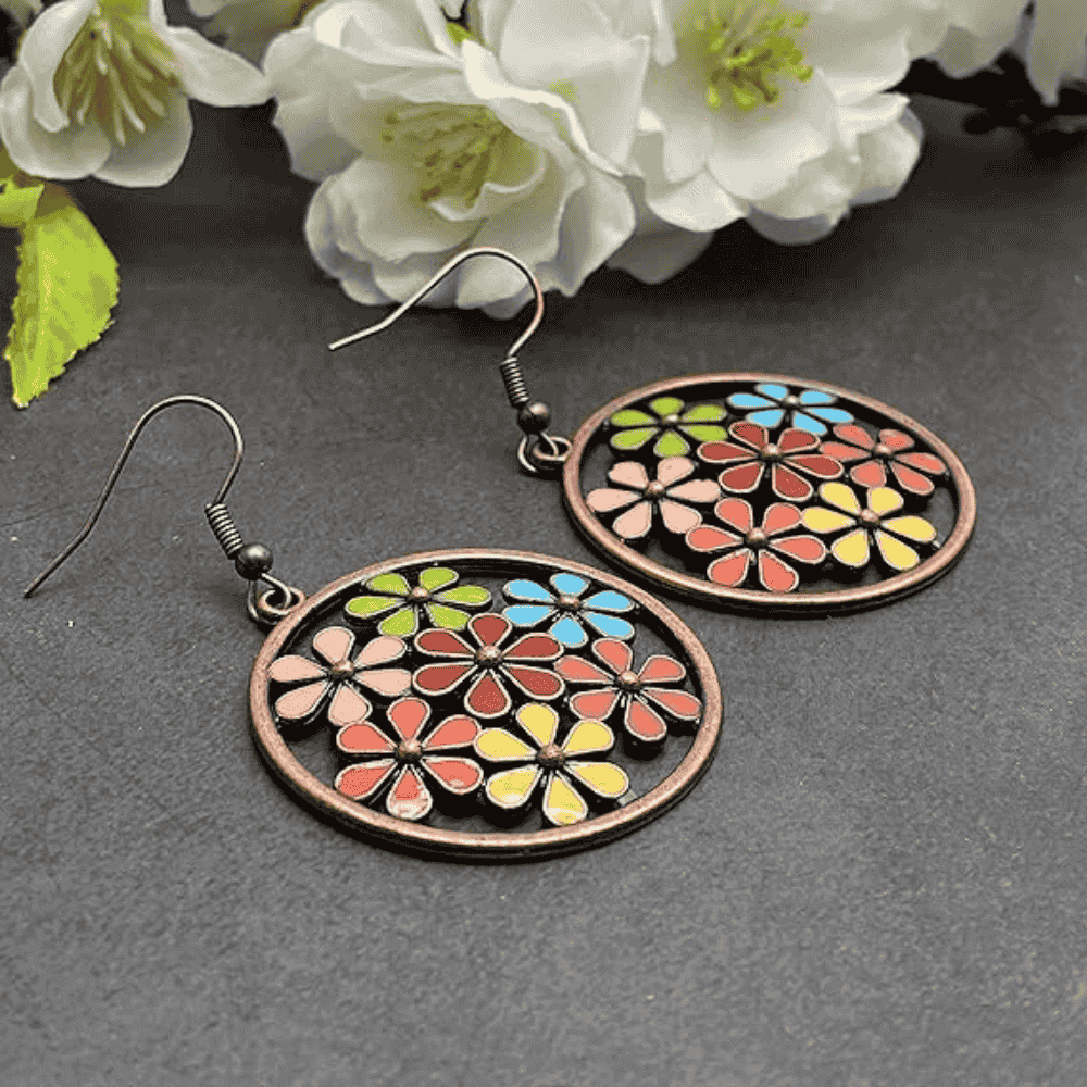 A pair of copper-tone boho earrings with multi-colored enamel flowers in a circular frame, resting on a dark surface with white flowers in the background.