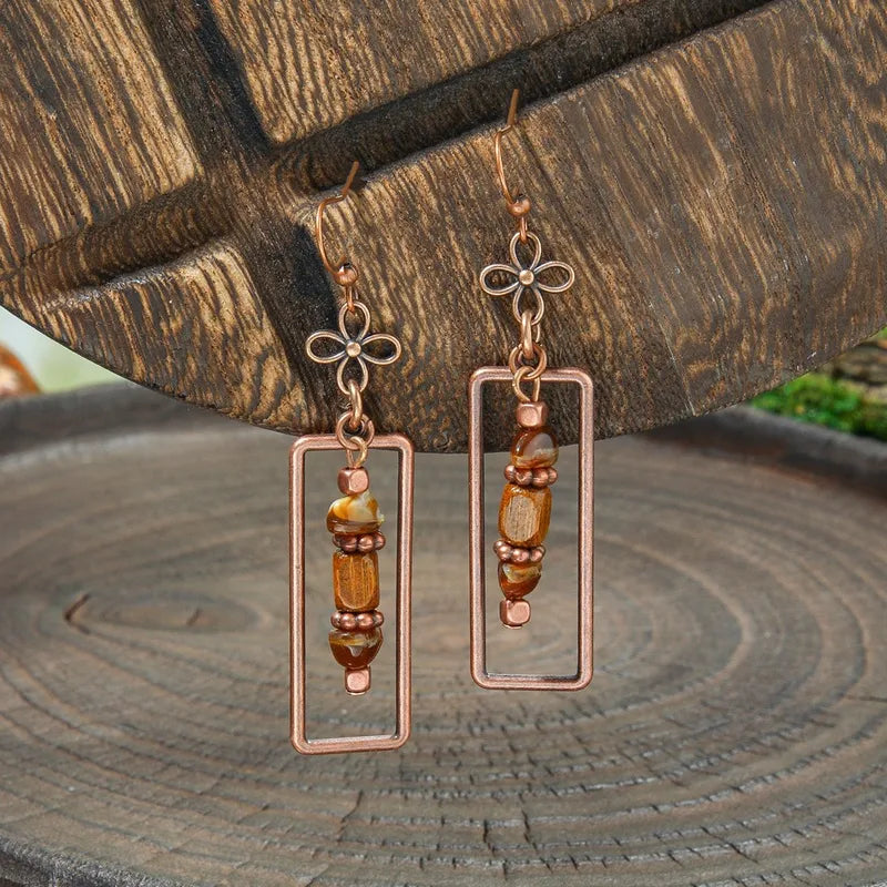A pair of boho earrings with copper rectangular frames and brown and gold beaded accents, hanging against a rustic wooden background.