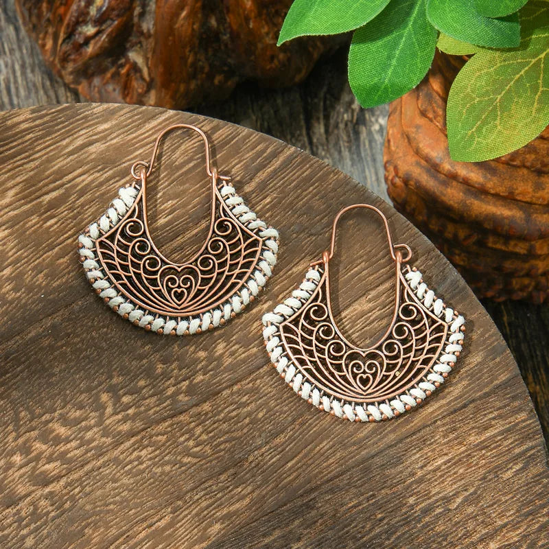 A pair of boho earrings featuring copper-toned filigree in a fan shape, adorned with white thread detailing along the edges, displayed on a wooden surface.