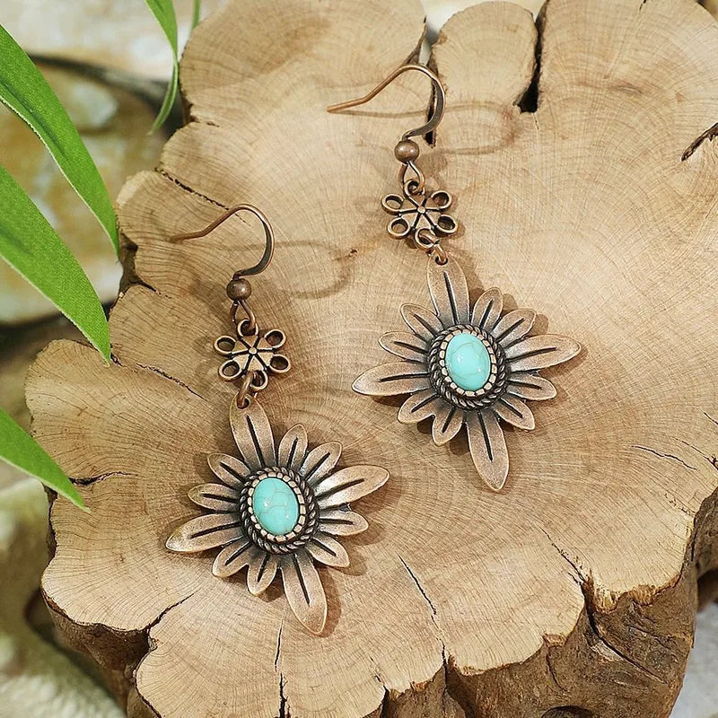 A pair of boho oxidised floral dangle earrings with turquoise stones, displayed on a wooden surface with green leaves in the background.