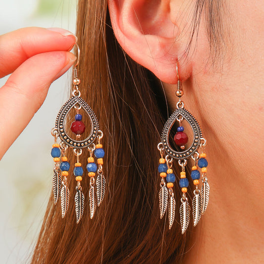 A woman wearing a pair of boho earrings with oxidised teardrop frames, blue and orange beads, and feather dangles, showcasing their size and style.