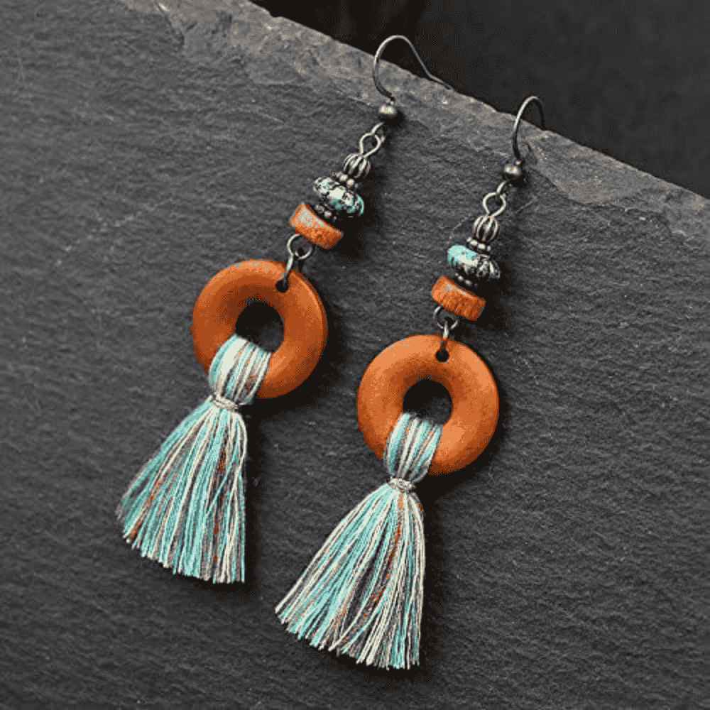 A pair of boho earrings with wooden hoops and blue, brown, and white tassels, laid on a dark textured surface.