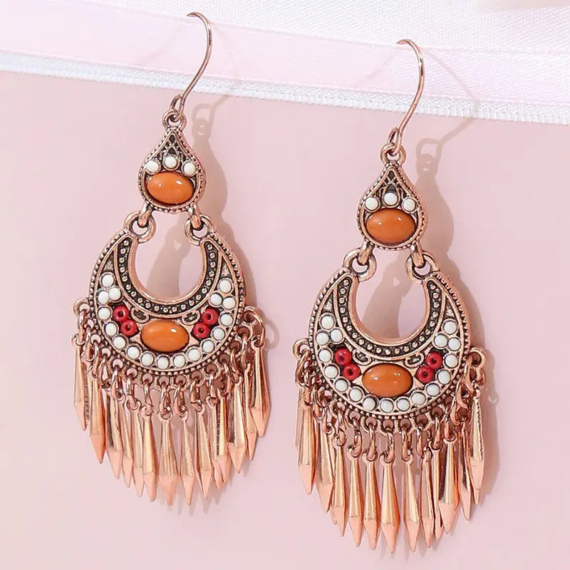 A pair of oxidised earrings with orange stones and tassels, hanging from a white ribbon against a light pink background.