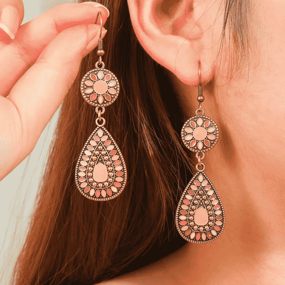A woman wearing a pair of long oxidised earrings with floral and teardrop designs in pink and white enamel.