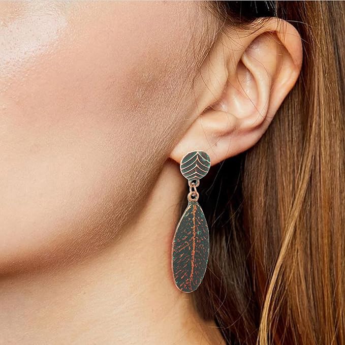 A close-up of a woman wearing a single oxidised earring with a copper-toned leaf drop and turquoise blue patina, featuring a chevron-patterned stud, against her ear.
