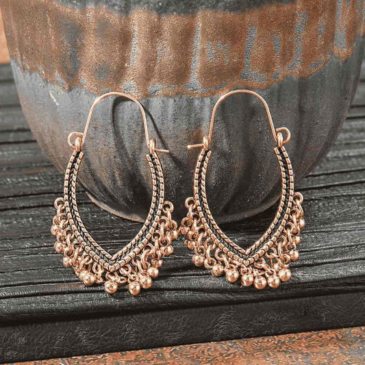 A pair of oxidised earrings with a ghungroo jingle design, showcasing an antique bronze finish and black detailing, set against a dark, textured pot and wooden surface.