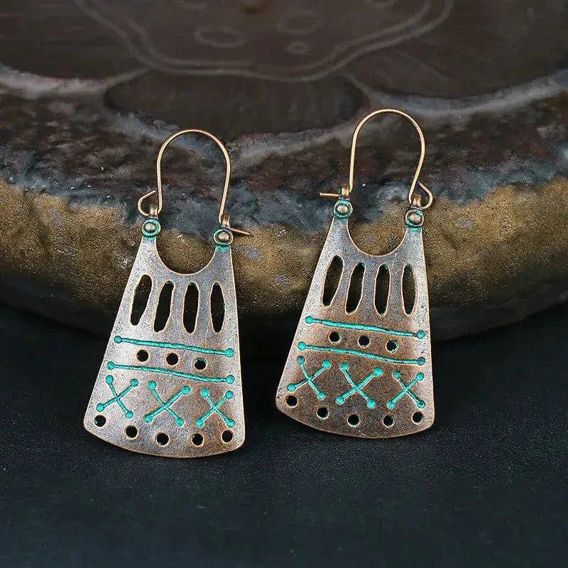 A pair of oxidised earrings featuring a tribal geometric pattern in bronze and teal, displayed on a dark surface with a textured brown rock in the background.