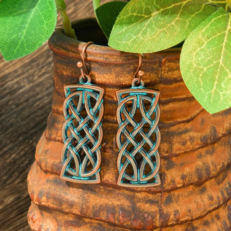 A pair of oxidised earrings with a Celtic knot design and blue patina, hanging in front of a rustic pot with green leaves.