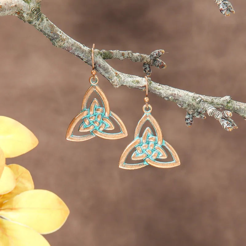 A pair of oxidised earrings with a Celtic knot design and blue patina, hanging from a tree branch with yellow leaves in the foreground.