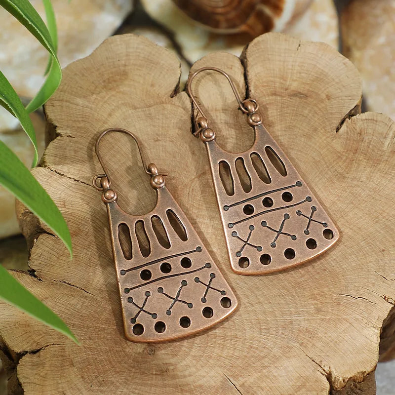 A pair of oxidised earrings with a geometric tribal motif, made of copper, resting on a wooden surface with green leaves in the foreground.