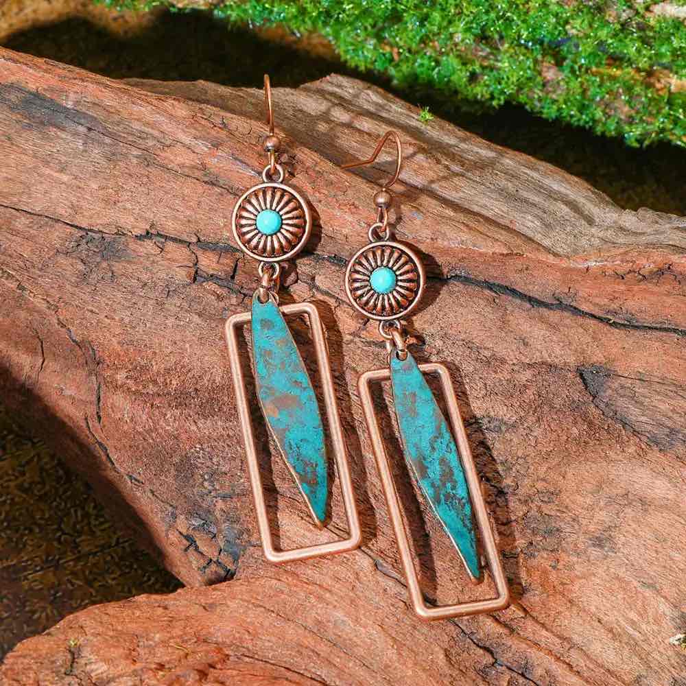 A pair of oxidised earrings with copper-toned frames and blue-green patina inserts, featuring a turquoise inlay at the top, resting on a wooden surface.