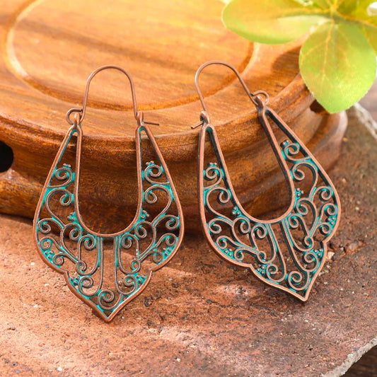 Two oxidised filigree drop earrings with a patina green accent, resting on a rustic wooden surface.