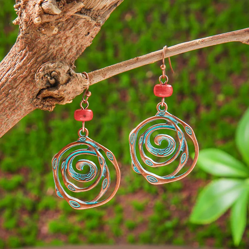 A pair of oxidised metal spiral drop earrings with copper and turquoise patina, hanging from a tree branch, against a green mossy background.