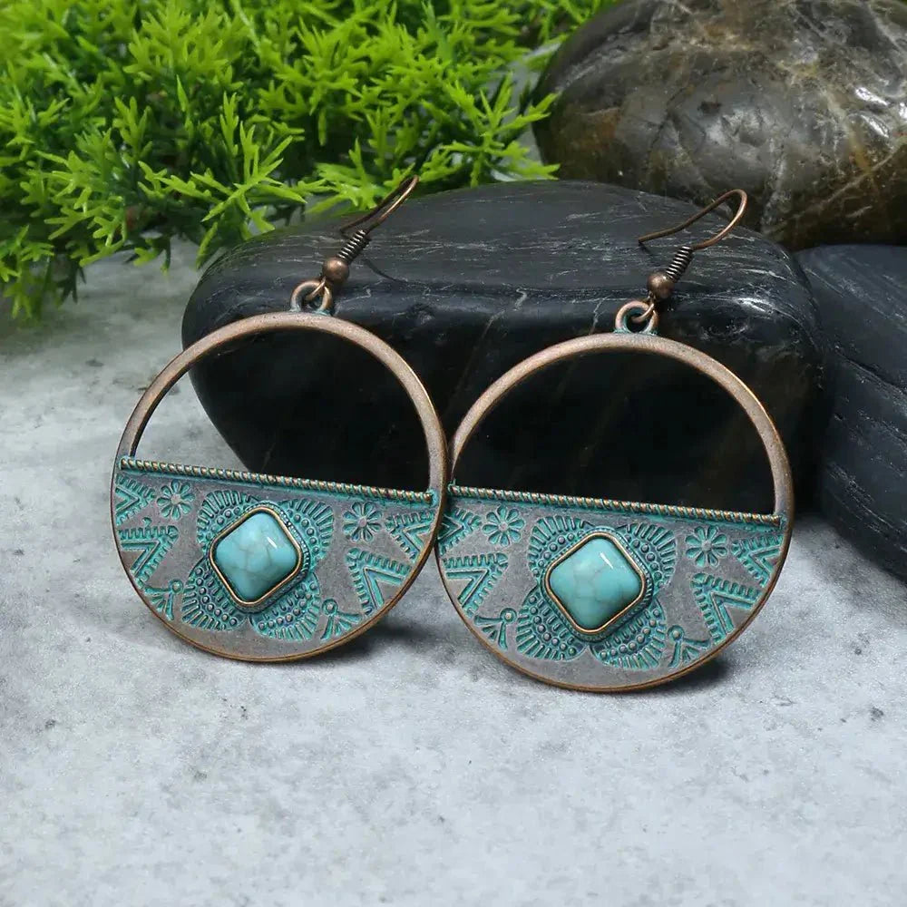 A pair of oxidised earrings with a half-hoop design and a turquoise stone, displayed on a grey stone surface with green foliage in the background.