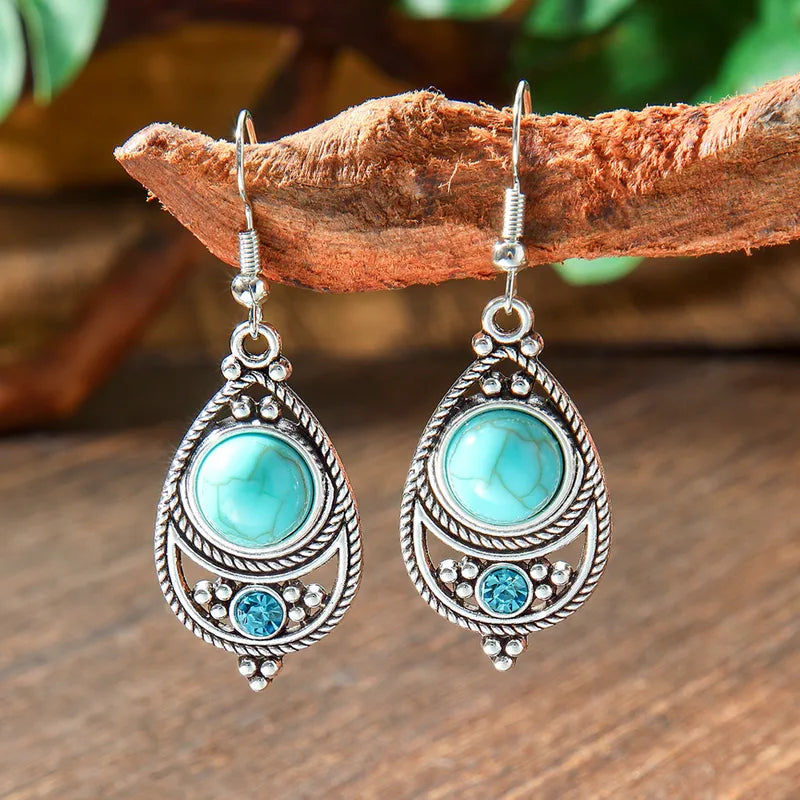A pair of oxidised earrings featuring a teardrop shape with a central turquoise gemstone and smaller blue rhinestones, hanging from a branch.