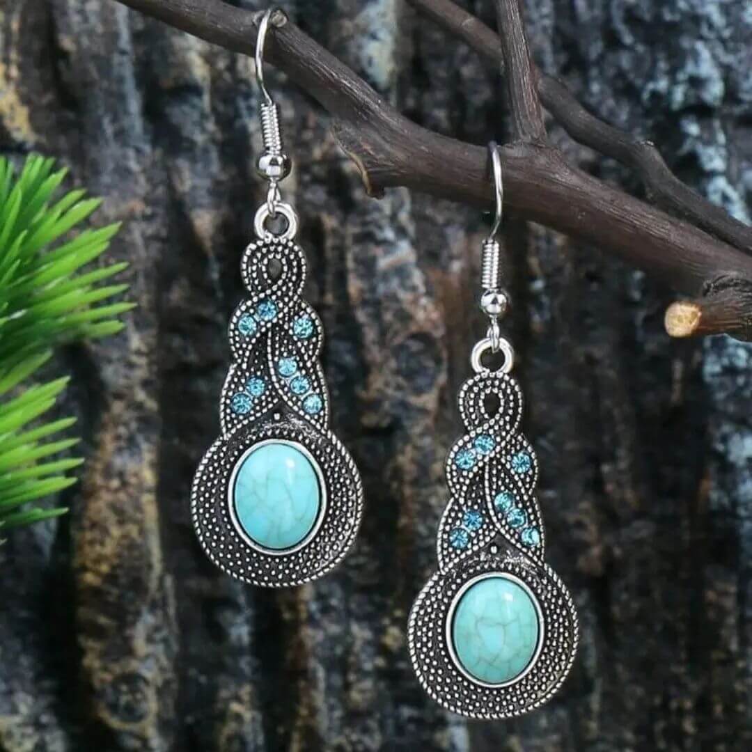 Two oxidised earrings with turquoise oval centers and sparkling blue accents, hanging from a rustic tree branch, showcasing their bohemian design.
