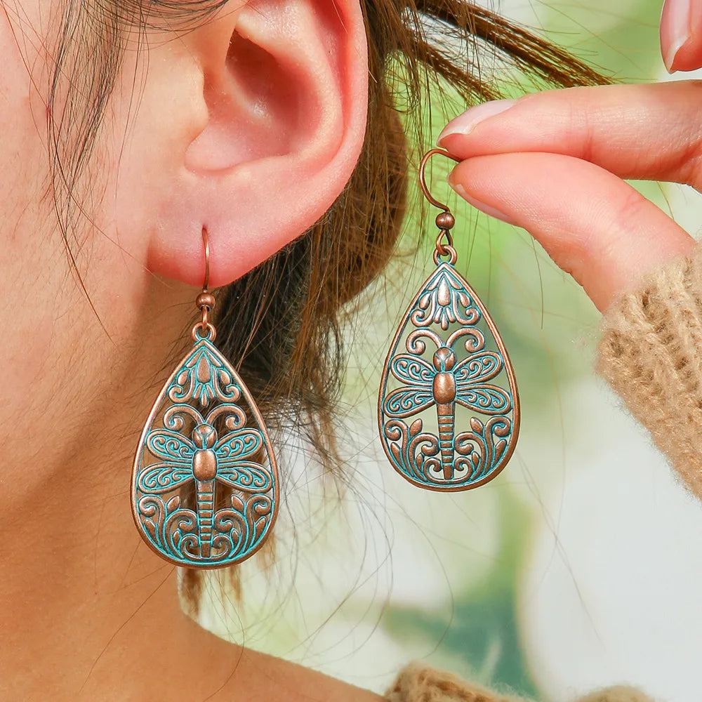 A woman wearing oxidised earrings with a teal dragonfly filigree design, held up to her ear.