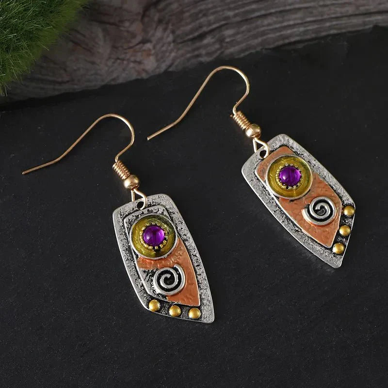 A pair of oxidised earrings with purple stones and tribal spiral designs, hanging from a branch against a blurred background.
