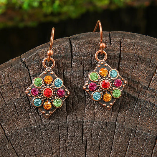 Close-up of a pair of bohemian earrings featuring a copper-toned rhombus shape with a central flower design made of colorful small stones, set against a dark wooden surface.