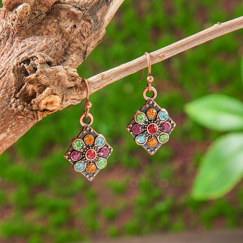 A pair of bohemian earrings with colorful stone flower designs, hanging from a tree branch with a blurred green background, showcasing their natural and earthy aesthetic.
