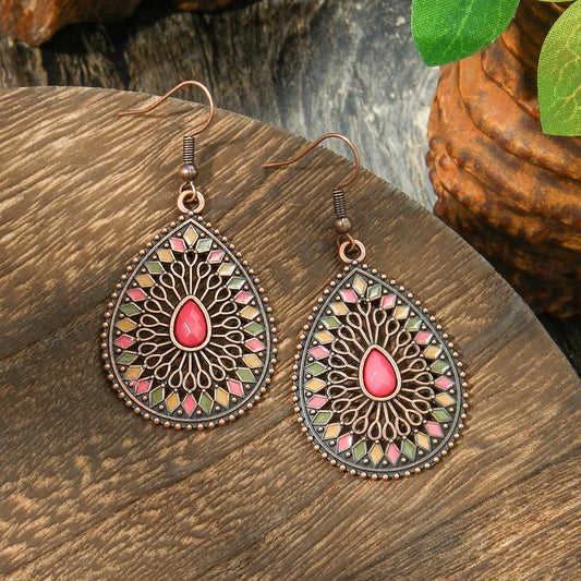 A pair of copper-toned boho earrings with a teardrop shape, featuring a colorful enamel geometric and filigree pattern around a central pink stone, displayed on a wooden surface.