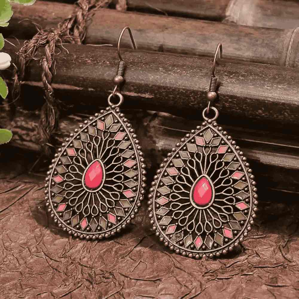 A pair of antique silver-toned boho earrings with a teardrop shape, showcasing a pink and grey enamel geometric and filigree design around a prominent pink stone, set against a rustic background.