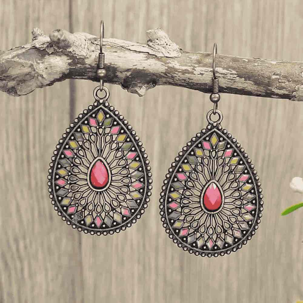 A pair of silver-toned boho earrings in a teardrop shape, adorned with a pastel pink, yellow, and green enamel geometric and filigree pattern surrounding a central pink stone, hanging from a wooden branch.