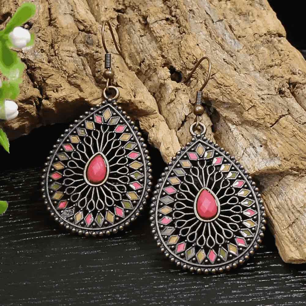 A close-up of a single copper-toned boho earring, highlighting its intricate filigree and geometric enamel work in shades of green, yellow, and pink, with a central pink stone, against a blurred wooden background.