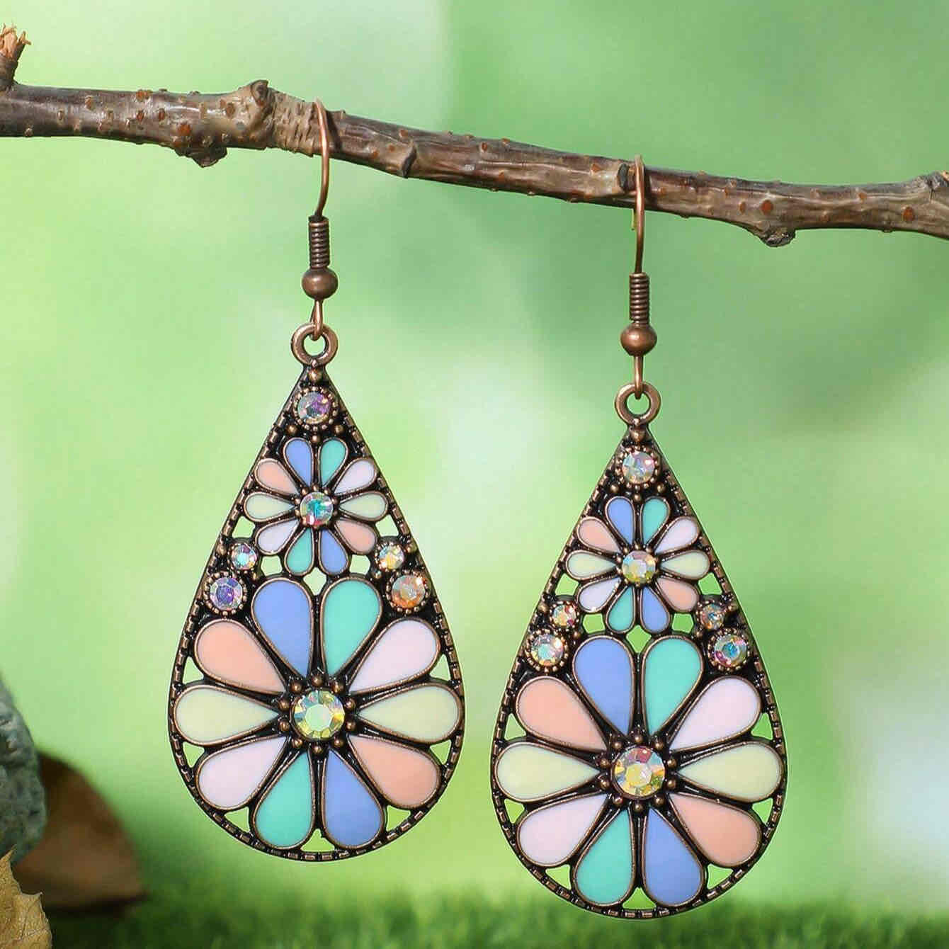 Pair of bohemian floral teardrop earrings with pastel enamel and small clear stones, hanging from a tree branch against a green background.