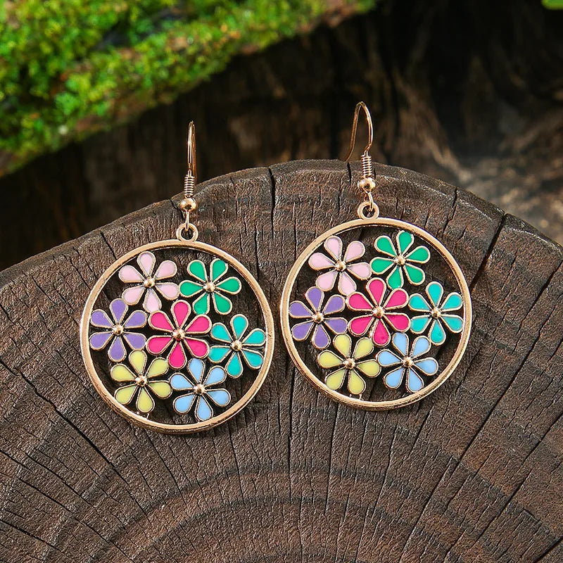 A pair of gold-tone boho earrings with multi-colored enamel flowers arranged in a circular frame, resting on a wooden surface.
