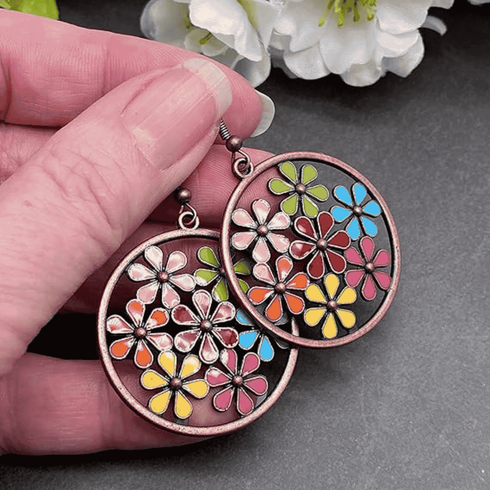 A hand holding a pair of copper-tone boho earrings with multi-colored enamel flowers in a circular frame, against a dark background.