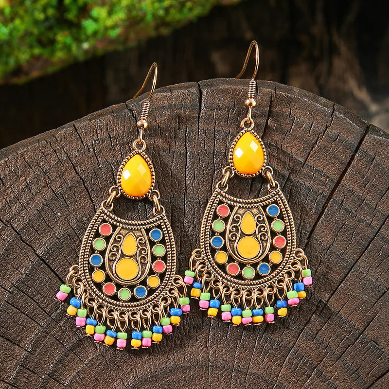 A pair of gold-toned boho earrings with yellow teardrop stones, colorful enamel details, and a vibrant beaded fringe, displayed on a rustic wooden surface.