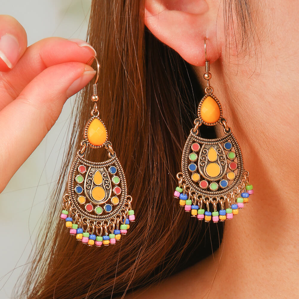 A model wearing a single gold-toned boho earring with a yellow teardrop stone, colorful enamel, and a beaded fringe, held gently by fingers against an ear.