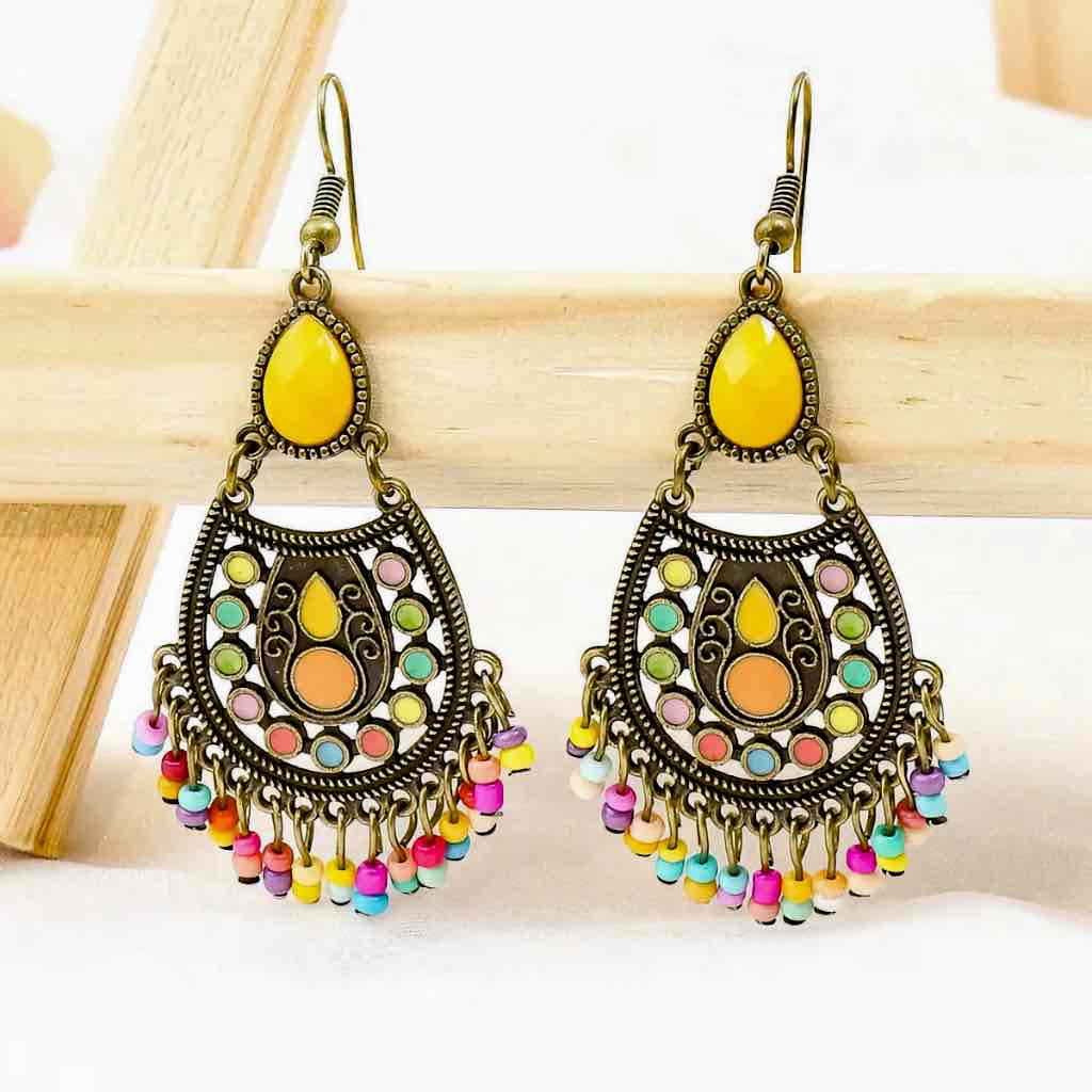 A pair of gold-toned boho earrings with yellow teardrop stones, colorful enamel accents, and a multicolor beaded fringe, displayed on a light wooden surface.