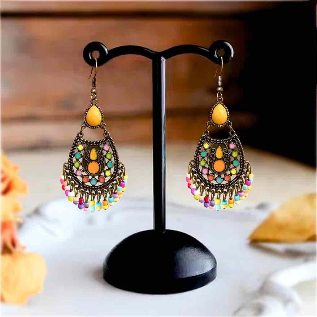 A close-up of a pair of gold-toned boho earrings featuring yellow teardrop stones, intricate colorful enamel work, and a lively beaded fringe, set against a blurred background.