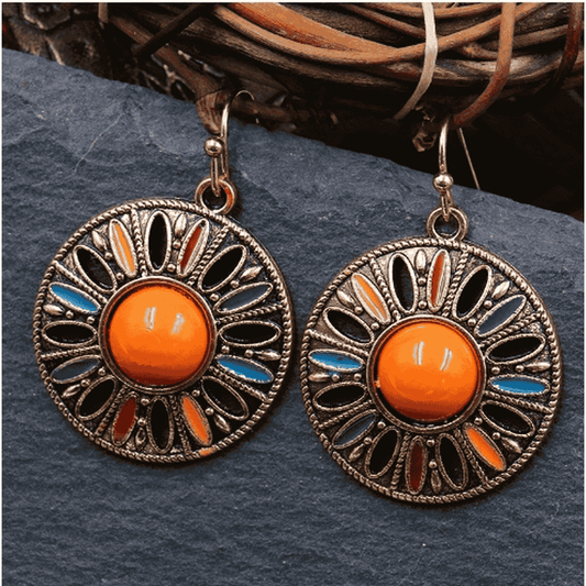 A pair of round boho earrings with a central orange stone and colorful enamel details, displayed on a dark slate surface.