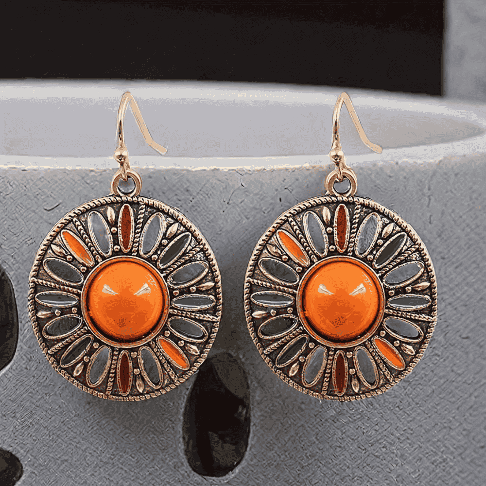 A pair of round boho earrings with a central orange stone and colorful enamel details, hanging against a light grey textured background.