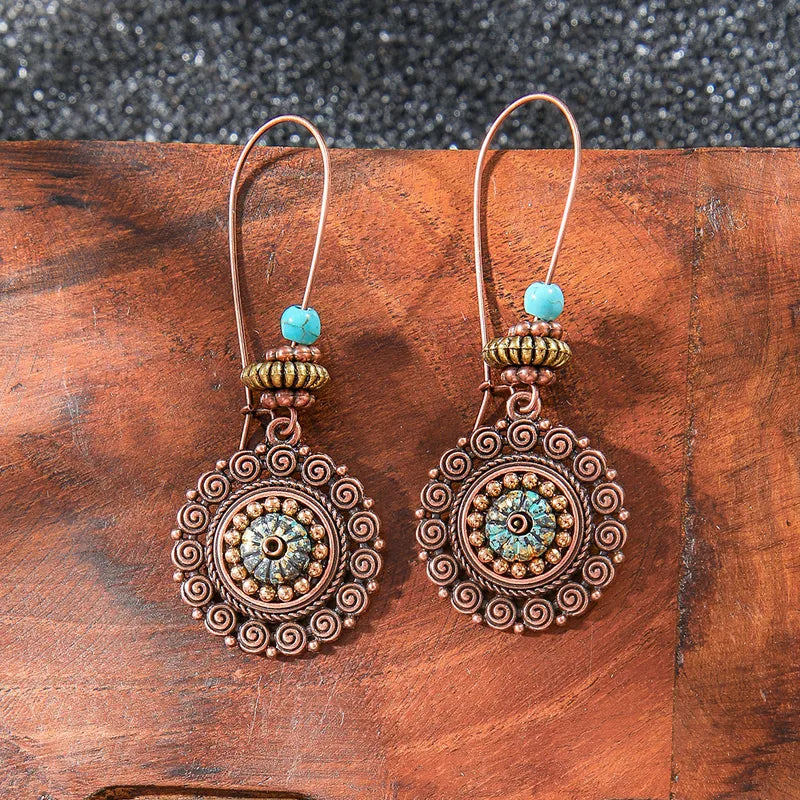 A pair of bohemian oxidised copper drop earrings with a central turquoise bead and intricate mandala-like filigree design, resting on a rustic wooden surface.