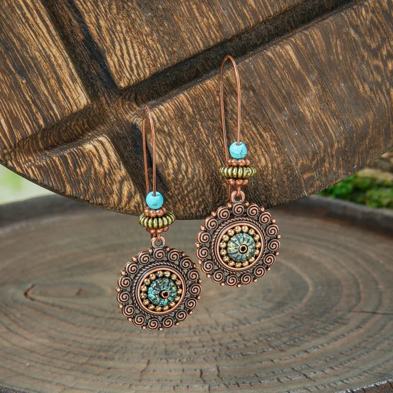 A pair of bohemian oxidised copper drop earrings featuring a turquoise bead and detailed filigree pattern, hanging against a textured wooden background.