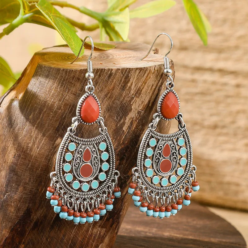 A pair of boho earrings featuring an oxidised silver-tone metal base with red teardrop stones, turquoise and red bead accents, and intricate tribal patterns, displayed on a wooden block.