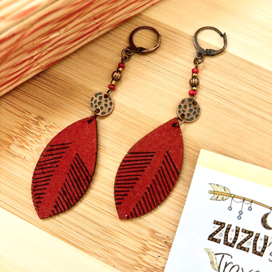 Two red leaf leather boho earrings with bronze and red bead accents, resting on a light wooden surface next to a small 'Earthy Origin' card.