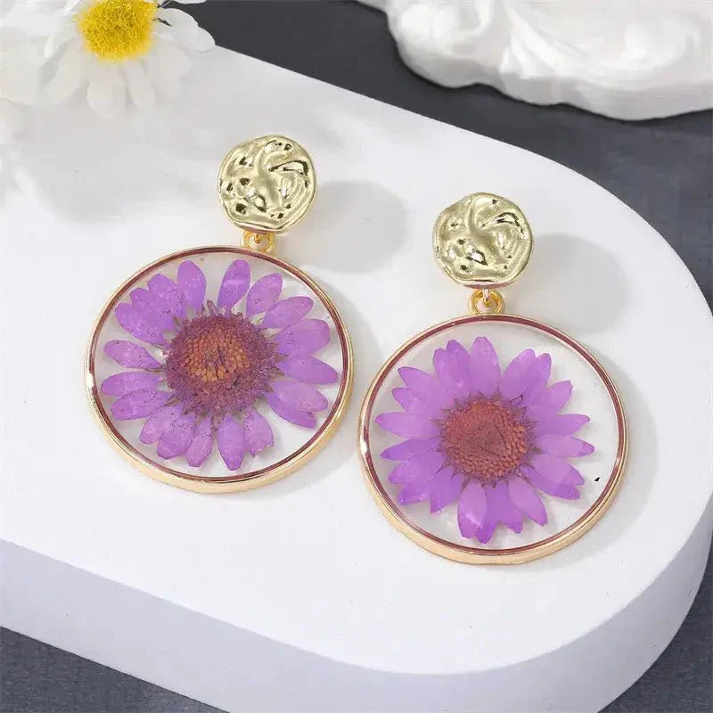 A pair of purple pressed flower boho earrings with gold-toned hammered studs, displayed on a white surface.