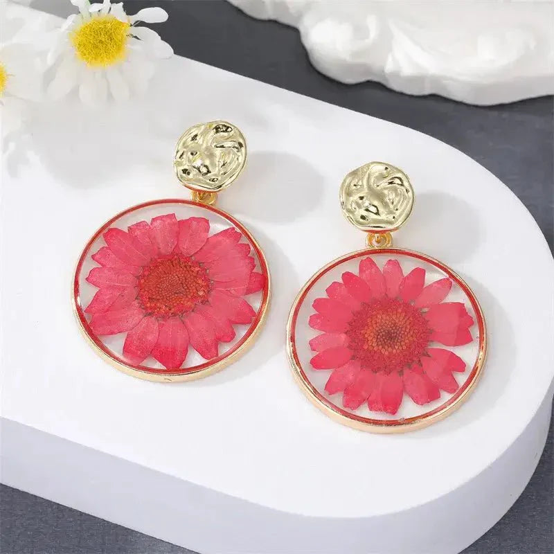 A pair of red pressed flower boho earrings with gold-toned hammered studs, displayed on a white surface.