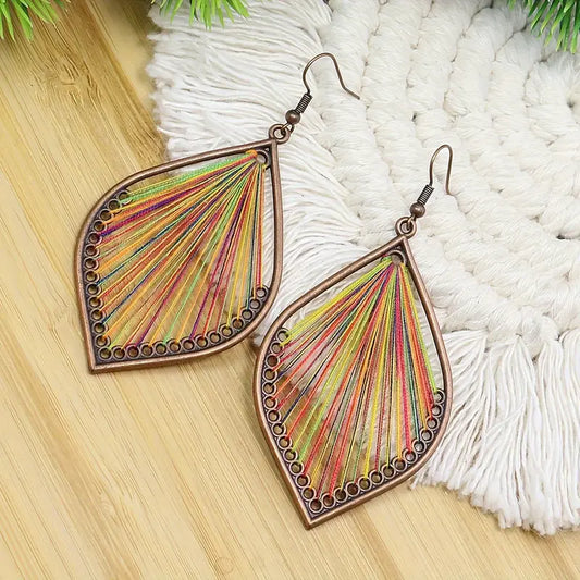 A pair of bohemian earrings with a bronze-toned teardrop frame filled with vibrant multicolor threads arranged in a fan pattern, resting on a white fringed mat.