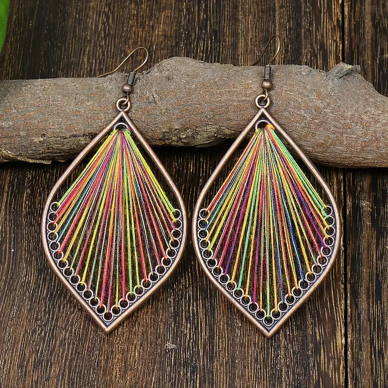 Two boho earrings, identical in design, featuring a bronze-toned teardrop frame with colorful threads, displayed against a dark wooden background.