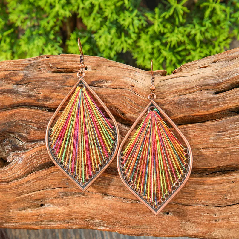 A pair of bohemian earrings showcasing a bronze-toned teardrop frame with a vibrant multicolor string art design, set against a rustic wooden log and green foliage.