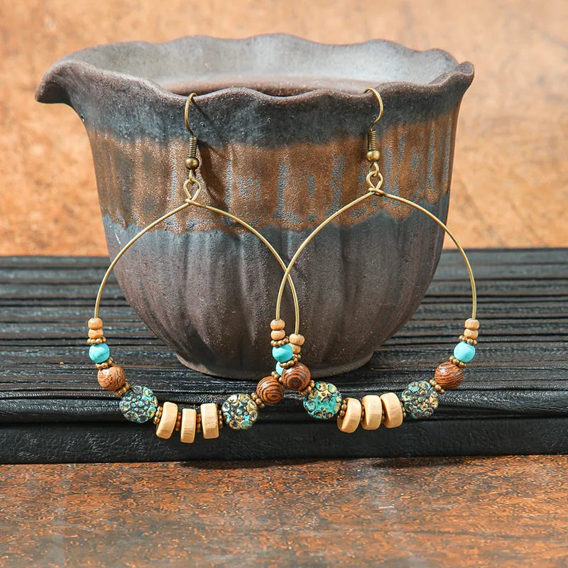 A pair of large, rustic boho earrings with a metal hoop, wooden beads, and turquoise accents, displayed against a dark ceramic pot.