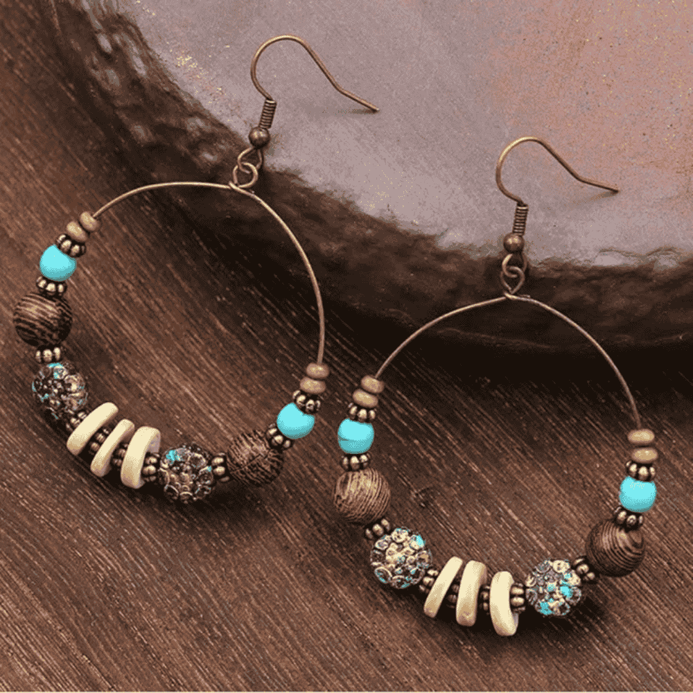 Close-up of a pair of bohemian earrings featuring wooden beads, patterned metal beads, and bright turquoise accents on a dark wooden surface.